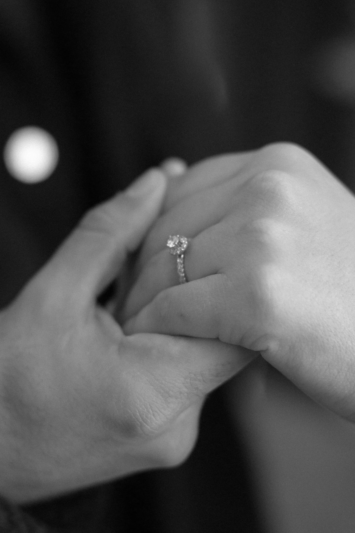 Two hands held together, one wearing a diamond engagement ring, photographed in black and white